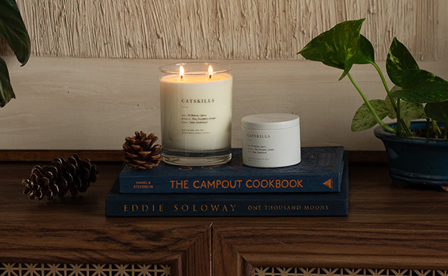 A lit white candle in a clear glass vessel and a small white jar, both labeled CATKILLS, displayed on a stack of blue books on a wooden surface with pinecones and a potted plant nearby.