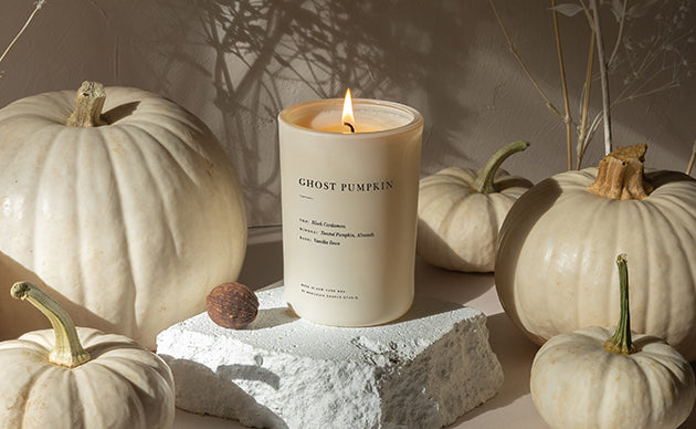 A lit cream-colored candle labeled 'GHOST PUMPKIN' sits on a white stone block, surrounded by decorative white pumpkins and a small brown acorn in a warm, styled setting.